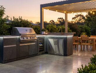 Modern outdoor kitchen with grill, sink, stone countertops and seating area in backyard.